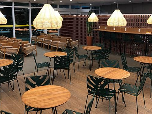 Imagens de diversas cadeiras de metal com formato de plantas e mesas redondas de madeira espalhadas em um espaço grande.